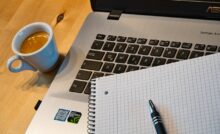 Part of a laptop, a notebook, pen and cup of tea on table background